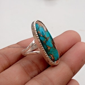 May include: A silver ring with an elongated oval turquoise and copper stone. The stone has a mix of blue-green and brown colors. The ring is held in a hand against a white background. The text "OMSAIJEWELRYCRAFT" is visible.