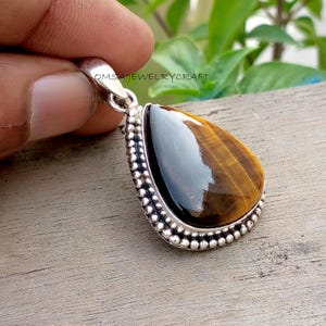 Puede incluir: Un colgante de piedra ojo de tigre en forma de lágrima engastado en un marco de plata con un borde de cuentas. La piedra presenta bandas alternas de brillo chatoyante marrón dorado y oscuro. El asa de metal tiene grabado el texto OMSAJEWELRYCRAFT.