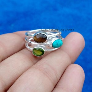May include: A stack of three silver rings, each featuring a single oval-shaped gemstone. The stones include a brown tiger's eye, a bright turquoise, and a translucent green peridot. The text OMSAIJEWELRYCRAFT is visible above the rings.