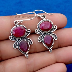 May include: A pair of silver earrings with oval and teardrop-shaped ruby gemstones. The earrings have a decorative silver border and a hook closure. The text "OMSAIJEWELRYCRAFT" is visible on the image.