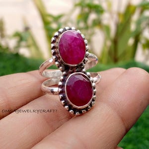 May include: A sterling silver ring featuring two oval-cut, deep red gemstones. The gemstones are set in ornate, beaded bezels. The ring has a split shank design and is displayed against a blurred green background.