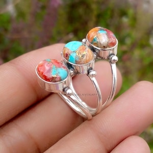 May include: Three silver rings with oval-shaped, multicolored stone settings. The stones feature a mix of red, turquoise, and gold hues. The rings are displayed on a hand, showcasing the jewelry's design and colors.