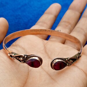 May include: A copper cuff bracelet featuring two teardrop-shaped dark red gemstones at the open ends. The metal band has a smooth finish, while the settings for the stones are decorated with twisted wire and intricate metalwork. Text reads OMSAIJEWELRYCRAFT.