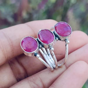 May include: A silver ring featuring three oval-cut, vibrant red gemstones. The ring has a unique design with a triple-band style, showcasing the polished gemstones. The setting appears to be detailed, adding to the ring's overall elegance.