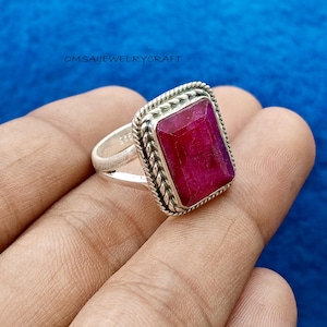 Op de afbeelding: Een zilveren ring met een grote, rechthoekige, dieprode edelsteen. De edelsteen is gezet in een decoratieve, gevlochten bezel. De ring heeft een eenvoudige, gladde band. De tekst "OMSAIJEWELRYCRAFT" is zichtbaar op de achtergrond.