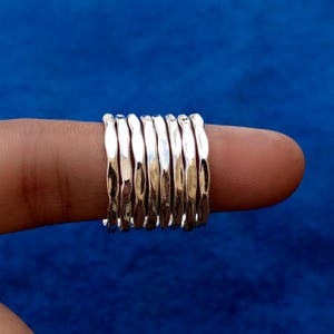 May include: A stack of silver rings with a hammered texture. The rings are displayed on a finger against a blue background. The rings have a shiny, metallic appearance.