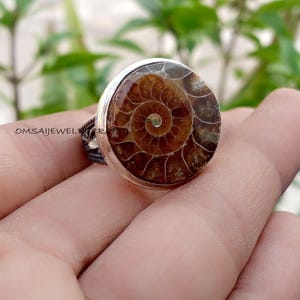 Könnte beinhalten: Ein Silberring mit einem kreisförmigen, polierten braunen Ammonitenfossil mit einem ausgeprägten Spiralmuster. Das Fossil ist in eine glatte Metallfassung eingefasst, und die Seite des Rings zeigt eine komplizierte, dunkle, texturierte Metallarbeit mit dem Text OMSAIJEWELRYCRAFT.