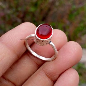 Könnte beinhalten: Ein silberner Ring mit einem großen, ovalen, tiefroten Edelstein. Das Ringband hat eine strukturierte, gehämmert wirkende Oberfläche. Der Edelstein ist in einer Zargenfassung gefasst. Der Ring wird in einer Hand gehalten, mit der Punze "925" sichtbar.