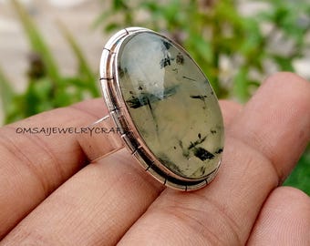 May include: A silver ring featuring a large, oval-shaped gemstone with a pale green hue and dark inclusions. The ring has a textured bezel and a simple band. The text "OMSAIJEWELRYCRAFT" is visible.