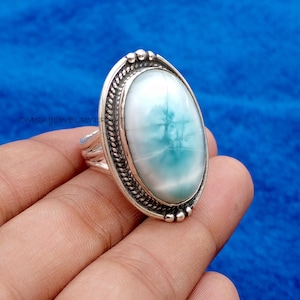 May include: Oval-shaped silver ring featuring a large, light blue gemstone with white and turquoise hues. The gemstone is set in a detailed silver band with a textured border. The ring is displayed against a blue background.