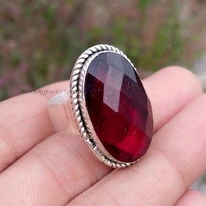 May include: A silver ring featuring a large, faceted, oval-shaped, deep red gemstone. The gemstone is set in a silver band with a twisted rope design around the stone. The ring is displayed on a hand.