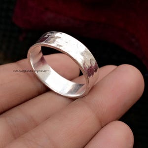 Könnte beinhalten: Ein silberner Ring mit gehammerter Textur. Der Ring hat ein schlichtes Banddesign mit leicht abgerundeter Innenseite und strukturierter Außenseite. Der Ring wird auf der Hand einer Person mit dunklem Hintergrund gezeigt.