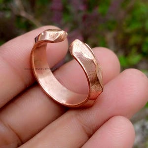 Può includere: Un anello aperto color rame con un design geometrico. L'anello ha una superficie strutturata e un gambo diviso. Il testo "OMSAJEWELRYCRAFT" è visibile sull'anello.