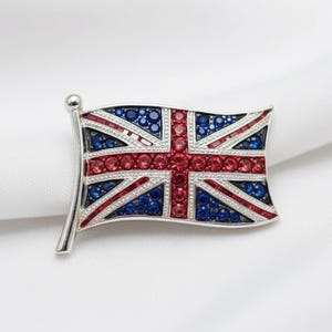 May include: A silver-toned brooch in the shape of the Union Jack flag. The flag design features red, white, and blue gemstones. The brooch has a silver pole and is set against a white background.
