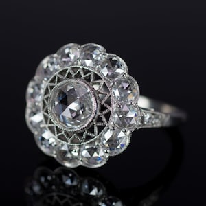 Könnte beinhalten: Ein silberner Ring mit einem großen, zentralen, klaren Edelstein, umgeben von kleineren, klaren Edelsteinen in einem floralen Muster. Das Band des Rings ist ebenfalls mit kleineren Edelsteinen verziert. Der Ring ist vor einem schwarzen Hintergrund platziert.