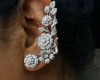 May include: A silver ear cuff featuring a floral design with multiple flower clusters and leaf accents. The ear cuff is encrusted with small, sparkling stones, creating a glamorous and elegant look. The design curves along the ear, with a separate flower cluster for the earlobe.