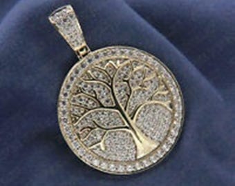 May include: A gold-toned pendant featuring a tree design, encrusted with small, sparkling stones. The pendant has a circular shape with a bail at the top for a chain. The tree's branches and trunk are visible within the circle.