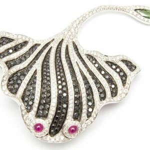 Op de afbeelding: Zilverkleurige broche in de vorm van een blad, versierd met zwarte en witte edelstenen. Twee rode edelstenen zijn aan de basis geplaatst en een groene edelsteen aan de punt. Het ontwerp is ingewikkeld en elegant.