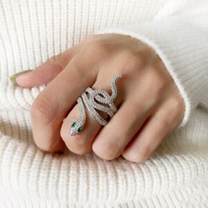 May include: A silver snake ring, adorned with clear crystals, coiled around a finger. The snake's head has two small green gems. The ring's design is intricate, with the crystals catching the light, creating a sparkling effect.