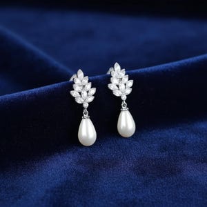May include: A pair of silver-toned earrings with a leaf-like design, adorned with clear crystals. Each earring features a pear-shaped, white pearl drop. The earrings are displayed against a dark blue velvet background.