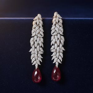 Puede incluir: Un par de elegantes pendientes colgantes. Cada pendiente presenta una cascada de pequeñas piedras brillantes con diseño de hojas, que culmina en una gran gema roja facetada en forma de lágrima. Sobre un fondo azul oscuro.