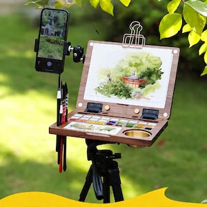 Puede incluir: Un kit de pintura de acuarela de madera con una paleta incorporada, una pequeña taza de latón y un soporte para teléfono inteligente. El kit está montado en un trípode negro, con una acuarela de un edificio en un paisaje verde.
