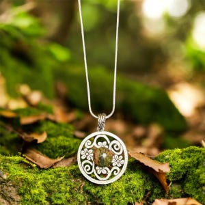 May include: A silver pendant necklace with a round, openwork design featuring floral and swirl patterns. The pendant holds a round, speckled green and brown stone. The necklace hangs against a backdrop of green moss and foliage.