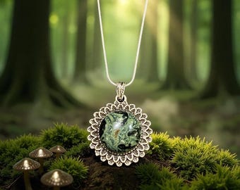 May include: A silver pendant necklace featuring a round, green stone with black inclusions, set in a decorative, scalloped frame. The pendant hangs from a delicate silver chain. The background is a blurred forest scene with moss and mushrooms.
