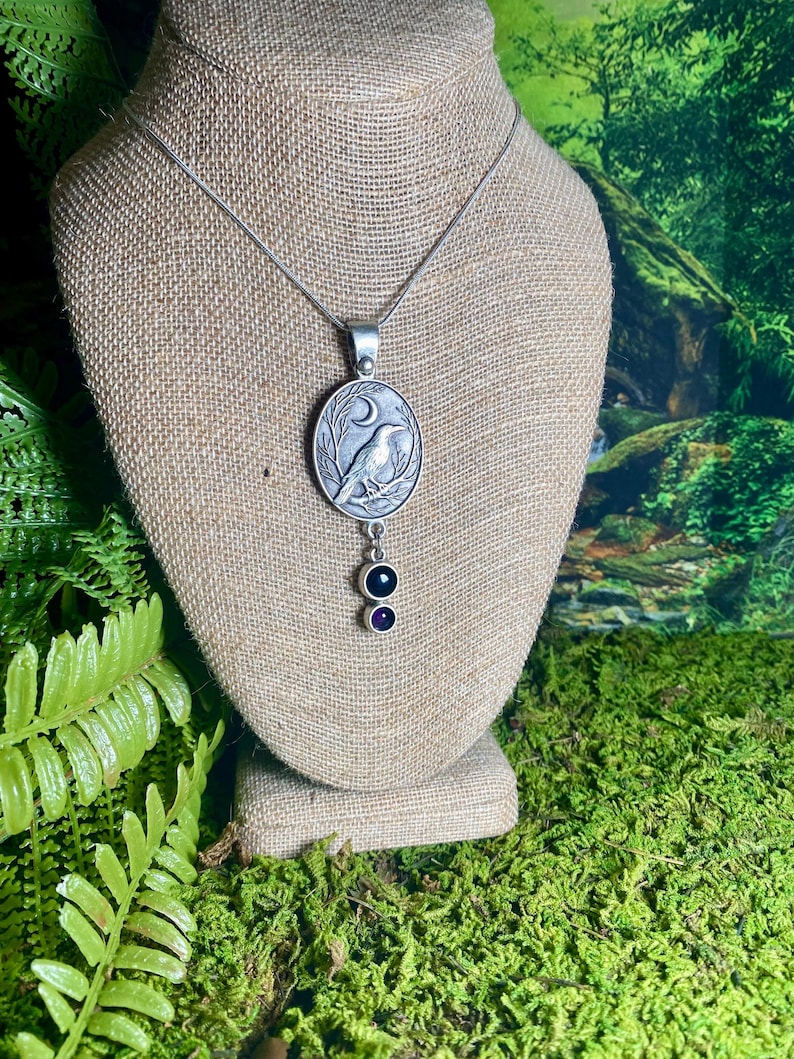 Pu&ograve; includere: Collana con pendente argentato con un design dettagliato di corvo all'interno di una cornice circolare, accentuato da una luna crescente. Il pendente &egrave; sospeso a una delicata catena e include due pietre preziose pendenti, una nera e una viola.
