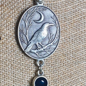 Pu&ograve; includere: Collana con pendente argentato. Il pendente ovale raffigura un corvo su un ramo sotto una luna crescente. Due gemme rotonde, blu scuro e viola, pendono sotto. La collana &egrave; su uno sfondo beige strutturato.