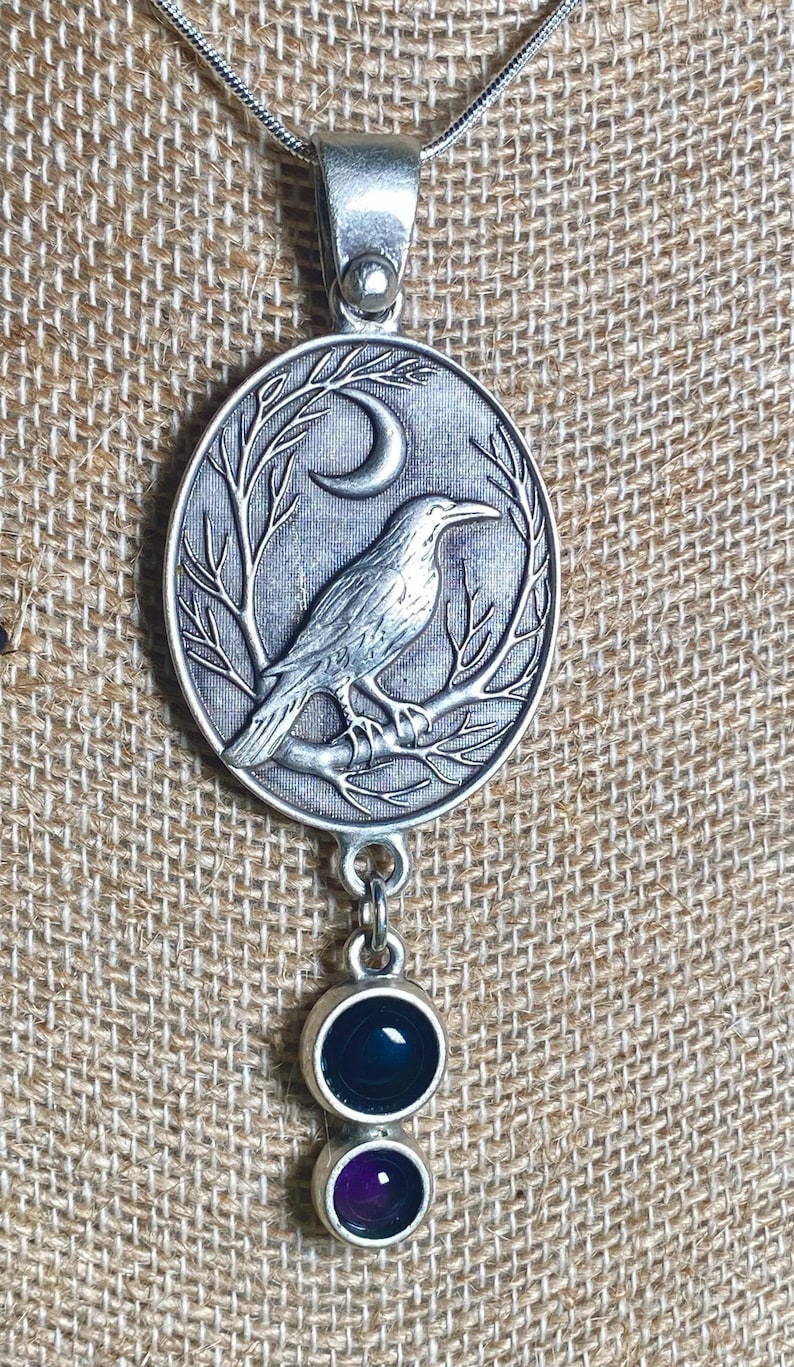 Pu&ograve; includere: Collana con pendente argentato. Il pendente ovale raffigura un corvo su un ramo sotto una luna crescente. Due gemme rotonde, blu scuro e viola, pendono sotto. La collana &egrave; su uno sfondo beige strutturato.