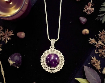 Puede incluir: Un collar con colgante de plata con una gema grande, redonda y de color púrpura oscuro. El colgante tiene un marco plateado adornado y festoneado. El collar se exhibe sobre una superficie negra, con elementos decorativos.