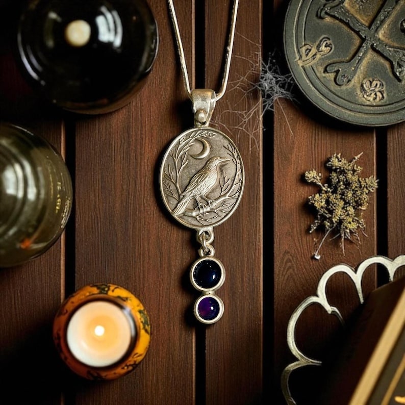 Pu&ograve; includere: Collana con pendente in argento raffigurante un corvo su un ramo sotto una luna crescente. Il pendente ha due gemme pendenti, una blu scuro e una viola. La collana &egrave; esposta su una superficie di legno con altri oggetti decorativi.