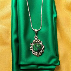 Peut inclure: Un collier en argent avec un pendentif orné d'une grosse pierre verte ronde avec des mouchetures sombres, sertie dans un cadre argenté décoratif en forme de fleur. Le collier est posé sur un fond de tissu vert et jaune.