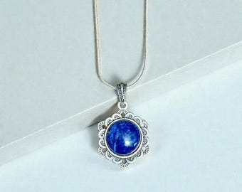 May include: A silver necklace with a round, deep blue pendant. The pendant is set in a decorative, openwork silver frame. The chain is a thin, silver snake chain. The necklace is displayed against a white background.