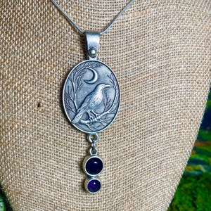 Pu&ograve; includere: Collana con pendente color argento con un pendente ovale raffigurante un corvo su un ramo sotto una luna crescente. Due gemme viola pendono sotto. La collana &egrave; esposta su una superficie con texture di iuta.