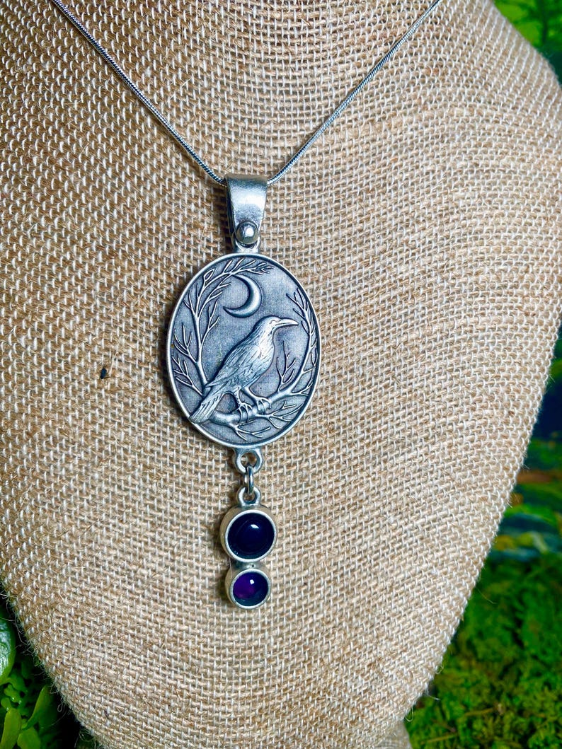 Pu&ograve; includere: Collana con pendente color argento con un pendente ovale raffigurante un corvo su un ramo sotto una luna crescente. Due gemme viola pendono sotto. La collana &egrave; esposta su una superficie con texture di iuta.