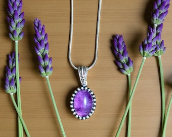 Puede incluir: Un collar de plata con un colgante ovalado morado, rodeado de pequeñas cuentas blancas. El colgante se encuentra sobre un fondo de ramitas de lavanda. El collar se exhibe sobre una superficie de madera.