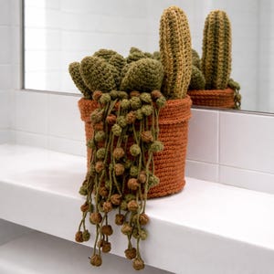 May include: A crocheted cactus arrangement in a terracotta-colored pot. The arrangement includes multiple green and brown cacti, with a trailing string of pearls plant. The pot is sitting on a white surface.