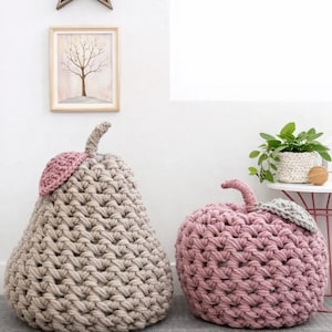 May include: Two large, handmade crochet fruit-shaped floor poufs. One is pear-shaped in beige with a pink leaf, the other is apple-shaped in pink with a gray leaf. They are on a gray rug.