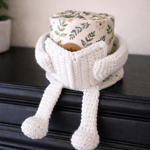 May include: A white, knitted toilet paper cozy shaped like a creature, with arms and legs. The cozy embraces a roll of toilet paper with a floral pattern. The cozy is sitting on a black surface.