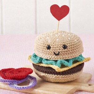 May include: A crocheted burger with a smiling face, a red heart on a toothpick, and additional crocheted burger components. The burger has a tan bun, a brown patty, and green lettuce. The burger is on a wooden cutting board.