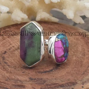 Puede incluir: Dos anillos de plata con engastes de piedras preciosas. Uno presenta una piedra verde y morada, el otro una piedra rosa y azul. Los anillos se exhiben sobre una superficie de madera.