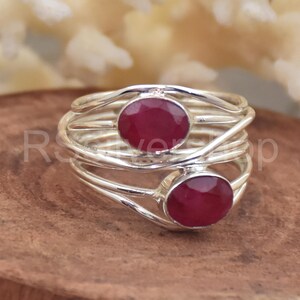May include: A silver ring featuring two oval, deep red gemstones. The ring's band has a woven design, and the gemstones are bezel-set. The ring is displayed on a wooden surface.