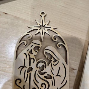 May include: A wooden Christmas ornament showing the Nativity scene. The ornament has a star at the top and an arched frame. The figures of Mary, Joseph, and baby Jesus are visible within the frame. The ornament is made of light-coloured wood.
