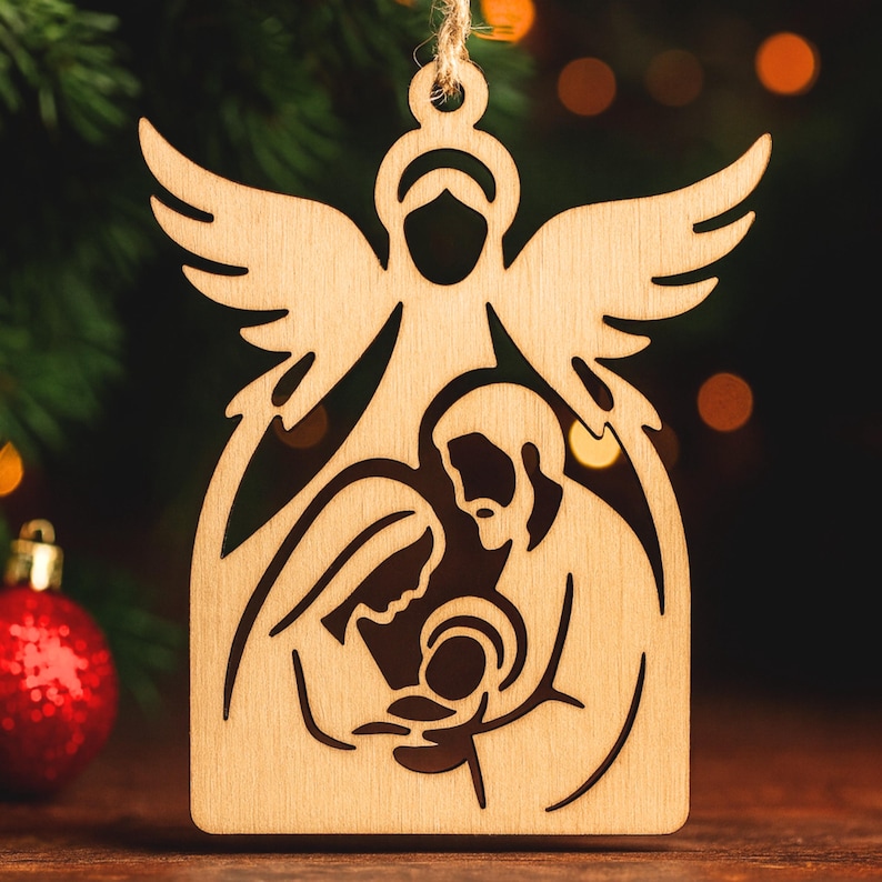 May include: A wooden Christmas decoration depicting an angel above the Holy Family. The ornament is crafted from light-coloured wood and hangs from a natural twine string. A red bauble is visible in the background, with a blurred festive backdrop.