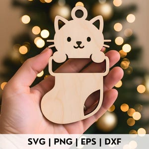 May include: A wooden Christmas ornament in the shape of a cat peeking out of a stocking. The ornament is made of light-colored wood and has a small hole at the top for hanging. The text "SVG | PNG | EPS | DXF" is at the bottom.