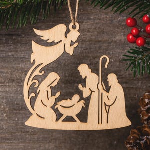 May include: Wooden Christmas decoration showing the Nativity scene. The ornament features an angel, Mary, Joseph, and baby Jesus in a manger. The ornament is made of light-coloured wood and is suspended from a twine string.