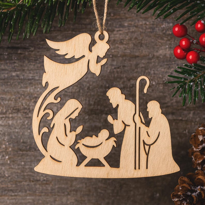 May include: Wooden Christmas decoration showing the Nativity scene. The ornament features an angel, Mary, Joseph, and baby Jesus in a manger. The ornament is made of light-coloured wood and is suspended from a twine string.