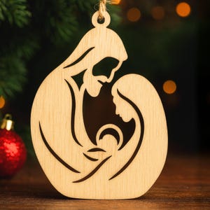 May include: Wooden Christmas ornament depicting the Holy Family: Joseph, Mary, and baby Jesus, in a cutout design. The ornament is made of light-coloured wood with a natural finish. A string is attached for hanging. The background includes a Christmas tree and bokeh lights.